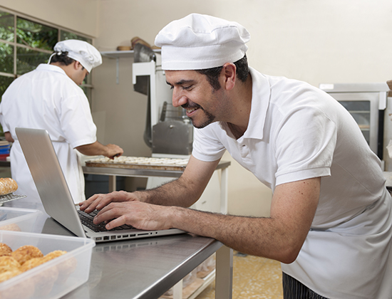 chef in white hat standing in kitchen and typing on a laptop computer