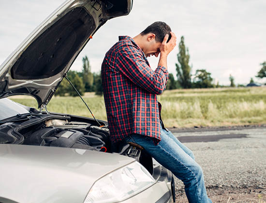 man in a flannel shirt and jeans, sitting on front of silver car with head in hands. car is broken down.